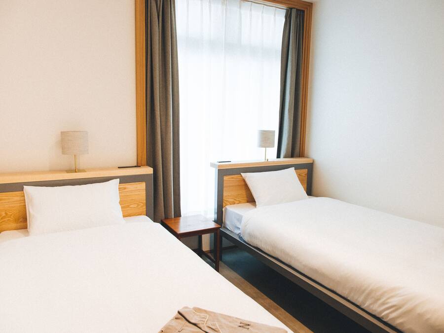 Dos camas individuales con cabeceros de madera, una mesita de noche con lámpara, y una ventana con cortinas.