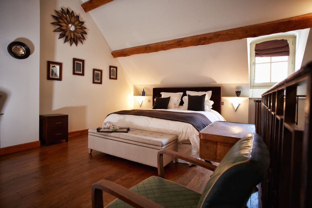 A bedroom with a large bed, wooden beams, a window with curtains, and framed pictures on the wall.