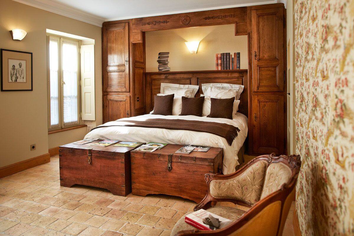 A bedroom with a large bed, wooden headboard, a chest of drawers, a chair, and a wall-mounted mirror.