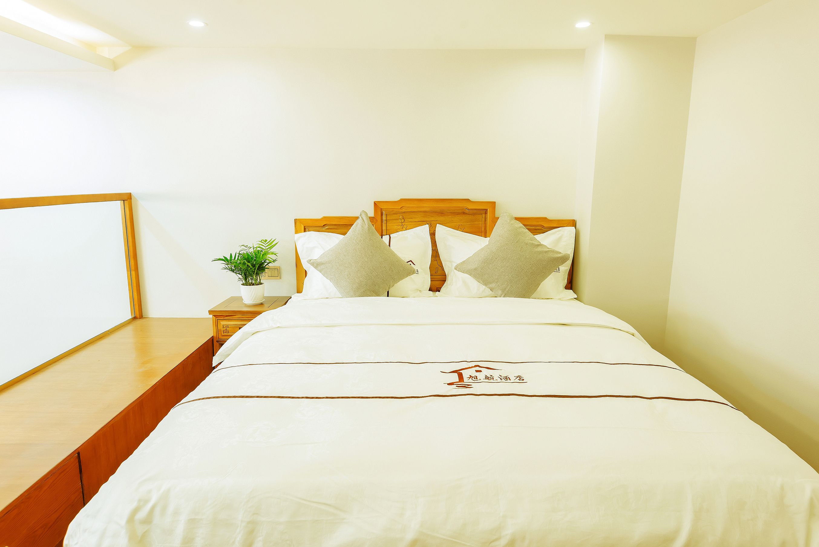 Una cama bien hecha con cabecera de madera, una mesita de noche con una planta y un espejo fijado a la pared.