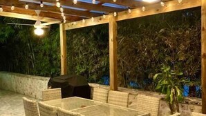 Een terras buiten met een glazen dak, rieten meubels en een barbecue.