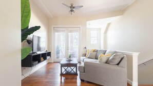 Ein Wohnzimmer mit grauem Sofa, Holzboden, Deckenventilator und Fernseher auf einem schwarzen Standfuß.