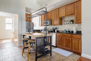 A kitchen with wooden cabinets, a stainless steel refrigerator, a microwave, and a sink. There is a wooden table with stools, and a black metal rack above the table.