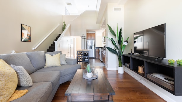 Ein modernes Wohnzimmer mit einem grauen Ecksofa, einem Holztisch, einem Flachbildfernseher auf einem schwarzen Standfuß und einer Treppe, die in ein Obergeschoss führt.