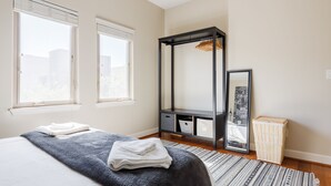 Ein Schlafzimmer mit einem Bett, zwei Fenstern, einem Kleiderschrank, einem Spiegel und einem kleinen Holzschemel.