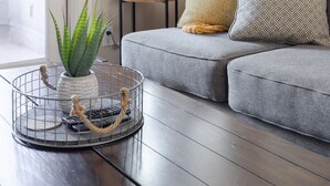 Ein Wohnzimmer mit grauem Sofa, einem kleinen Beistelltisch, einer Stehlampe und einem Topfpflanzen.