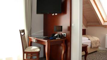 A hotel room with a wooden desk, a chair, a television mounted on the wall, a bedside table, and a bed with a patterned bedspread.