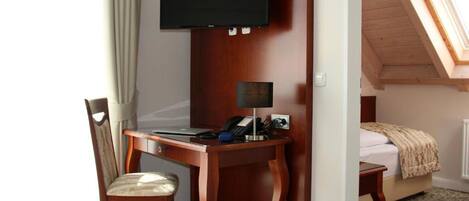 Ein Hotelzimmer mit einem Holztisch, einem Stuhl, einem an der Wand befestigten Fernseher, einem Nachttisch und einem Bett mit gemustertem Bettbezug.