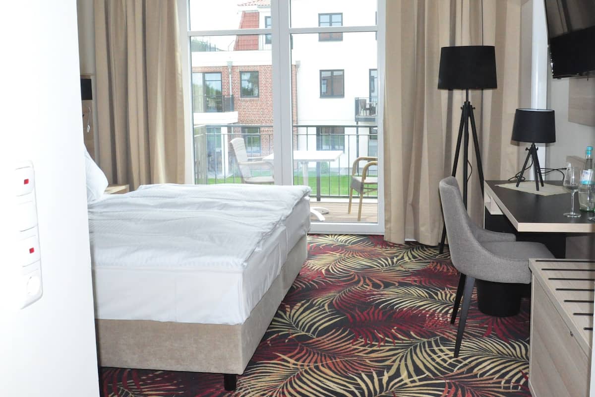 A hotel room with a bed, a desk with a lamp, a chair, and a balcony with a view of buildings.