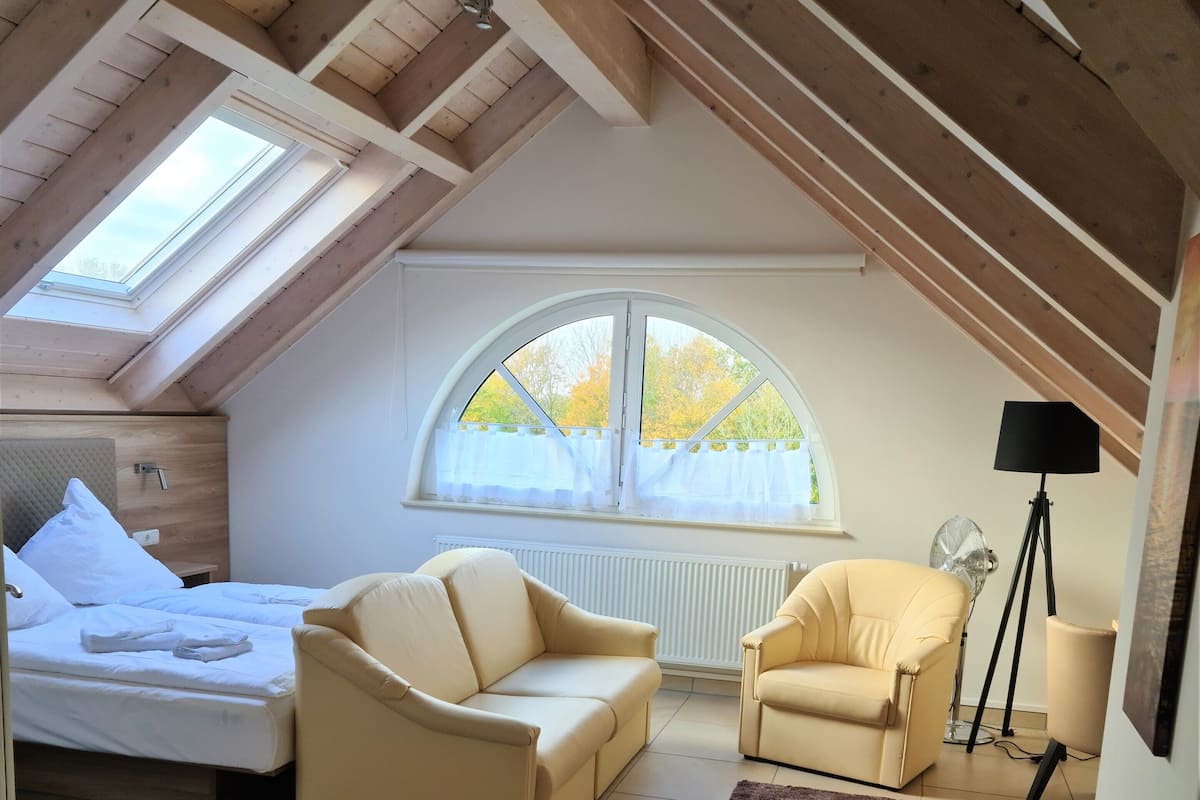 A cozy attic bedroom with a bed, two sofas, a floor lamp, and a large window.