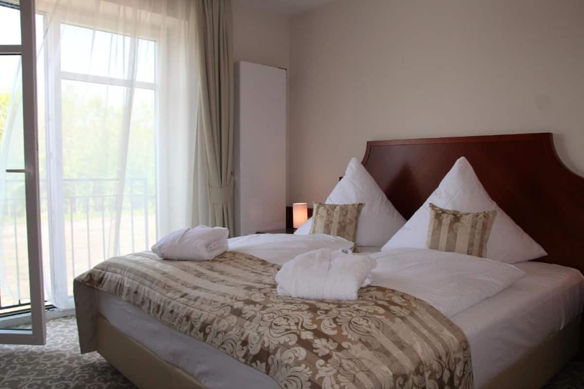 A hotel room with a large bed, two pillows, a wooden headboard, a window with curtains, and a door.