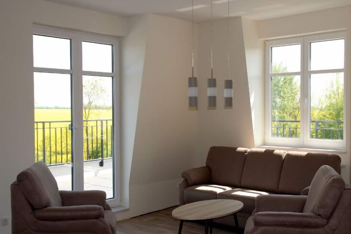 Un salon comprenant un canapé, deux fauteuils, une petite table ronde et de grandes fenêtres donnant sur un champ verdoyant.