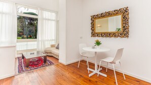 A modern living room with a wooden floor, a round white table with two chairs, a large ornate mirror, and a view of a garden through a large window with sheer curtains. - Maison Giulivia a San Pietro (Rome)