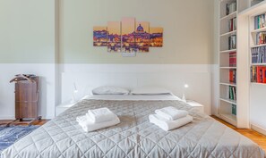A neatly made bed with a quilted cover, two rolled towels, a bedside table with a lamp, a wall-mounted painting, and a bookshelf filled with books. - Maison Giulivia a San Pietro (Rome)