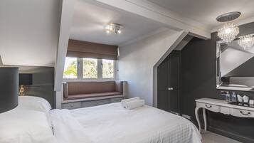 A bedroom with a large bed, a bench by the window, a vanity with a mirror, and a chandelier.