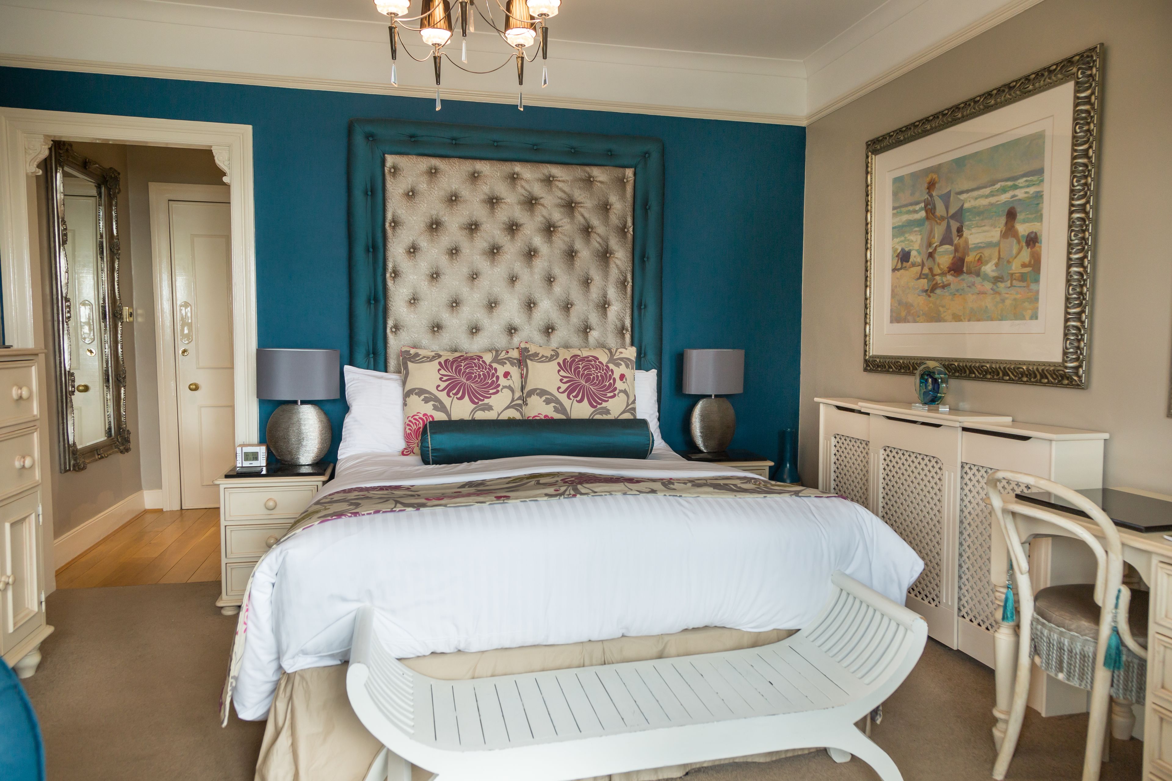 A bedroom with a large bed, two bedside tables with lamps, a dresser, a chair, and a framed painting on the wall.