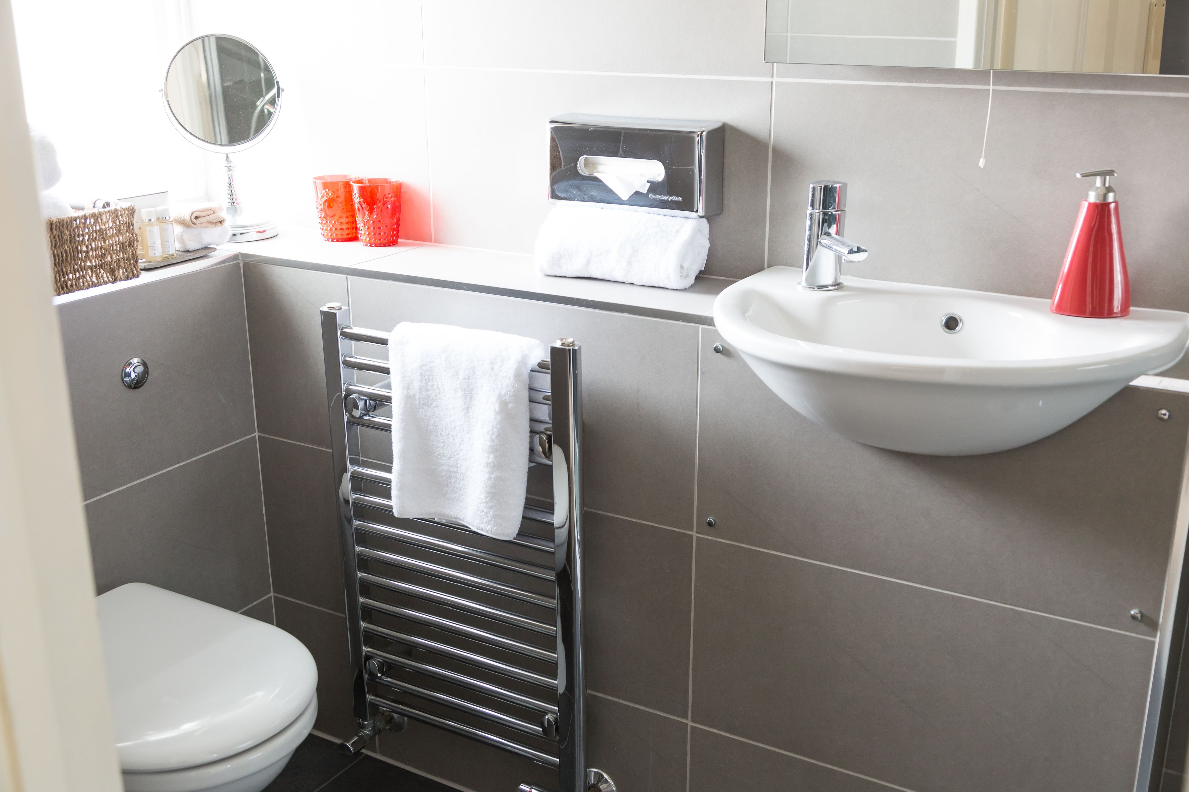 Ein Badezimmer mit einer weißen Toilette, einem Handtuchhalter mit einem weißen Handtuch, einem Waschbecken mit einem roten Seifenspender und einem Spiegel.
