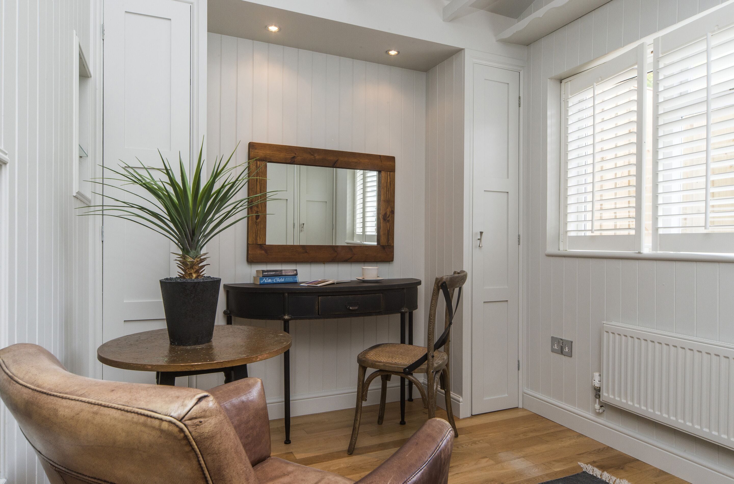 A room with a wooden dining table, two chairs, a potted plant, a mirror, a small desk, and a radiator.