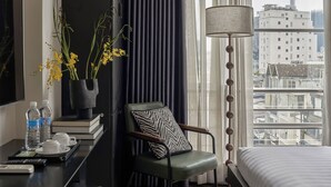 A modern hotel room with a bed, a desk with a lamp, a chair, a view of the city, and a vase with yellow flowers. - The One Premium Hotel (Ho Chi Minh City)