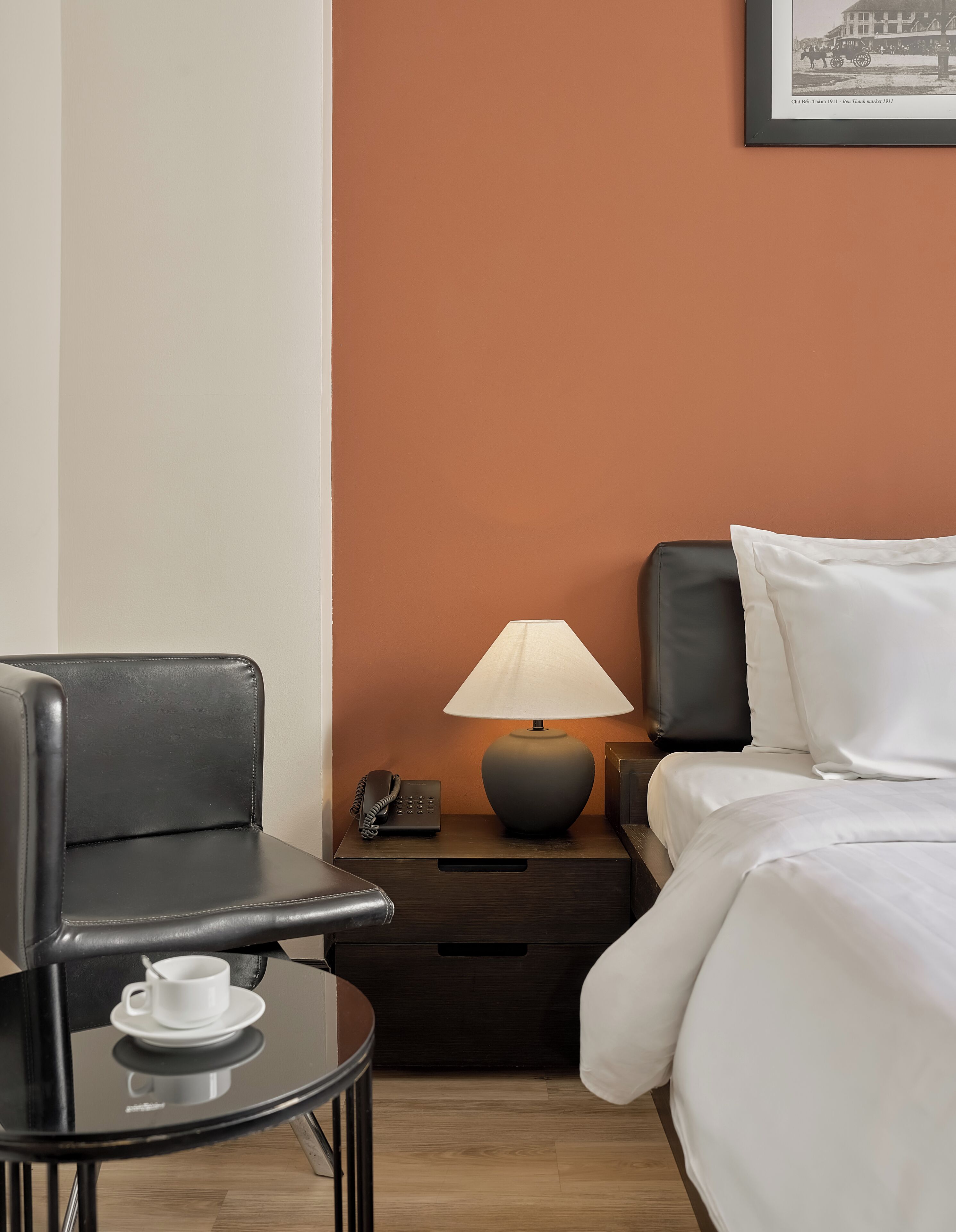 Habitación de hotel con una cama, una mesita de noche con lámpara, teléfono, silla y una pequeña mesa con una taza.