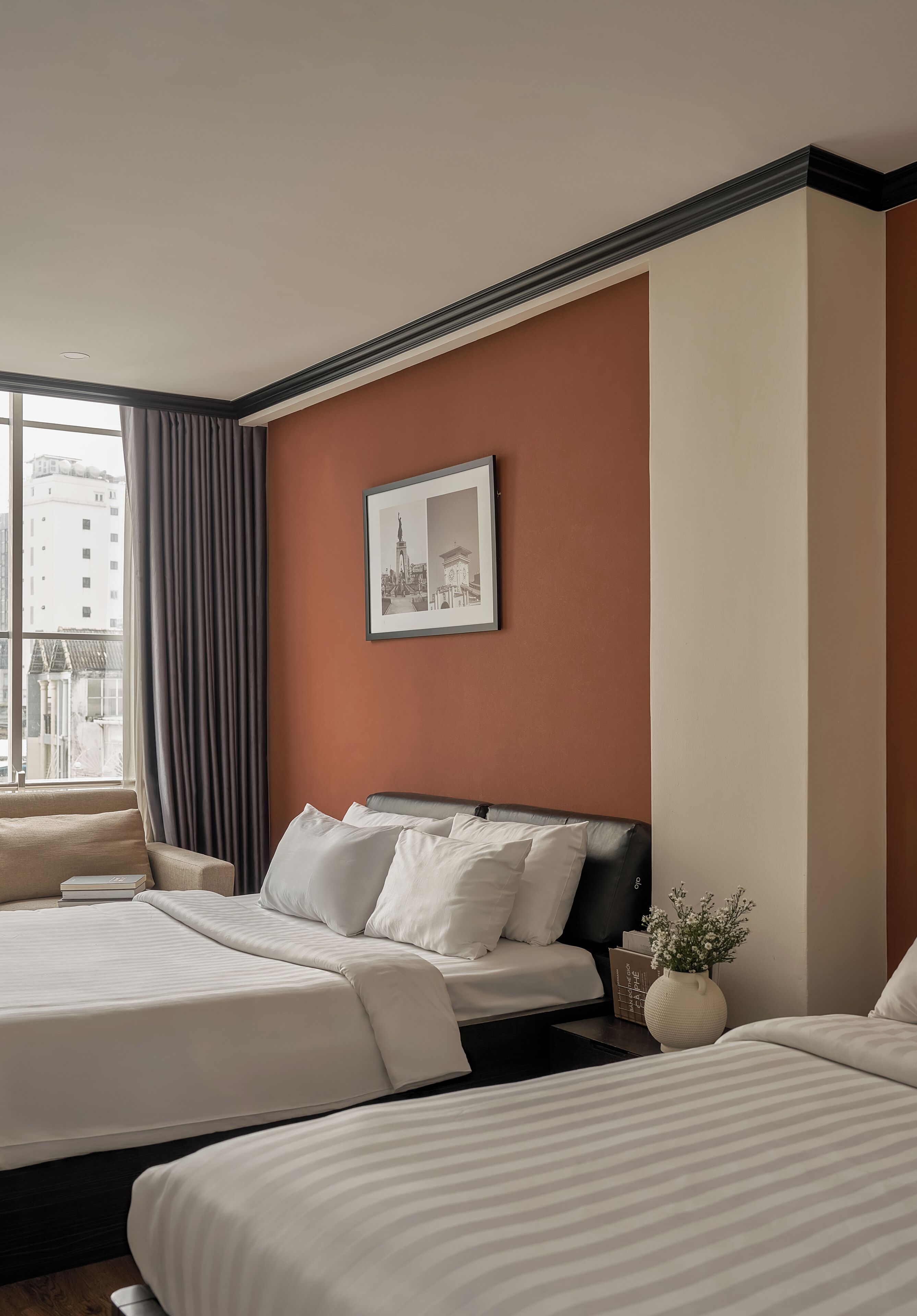 Ein Hotelzimmer mit einem großen Bett, einem kleineren Bett, einem Fenster mit Vorhängen, einem gerahmten Bild an der Wand und einer Vase mit Blumen.
