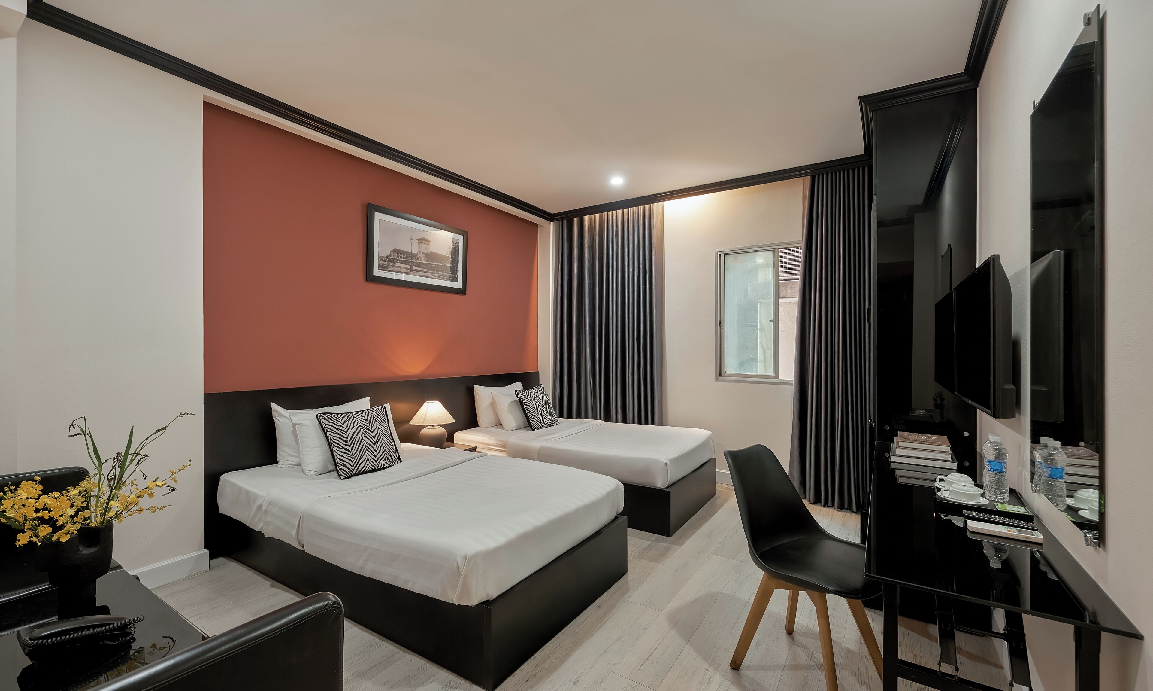 Ein Hotelzimmer mit zwei Betten, einem Schreibtisch mit Stuhl, einem Fernseher und einem Fenster mit Vorhängen.