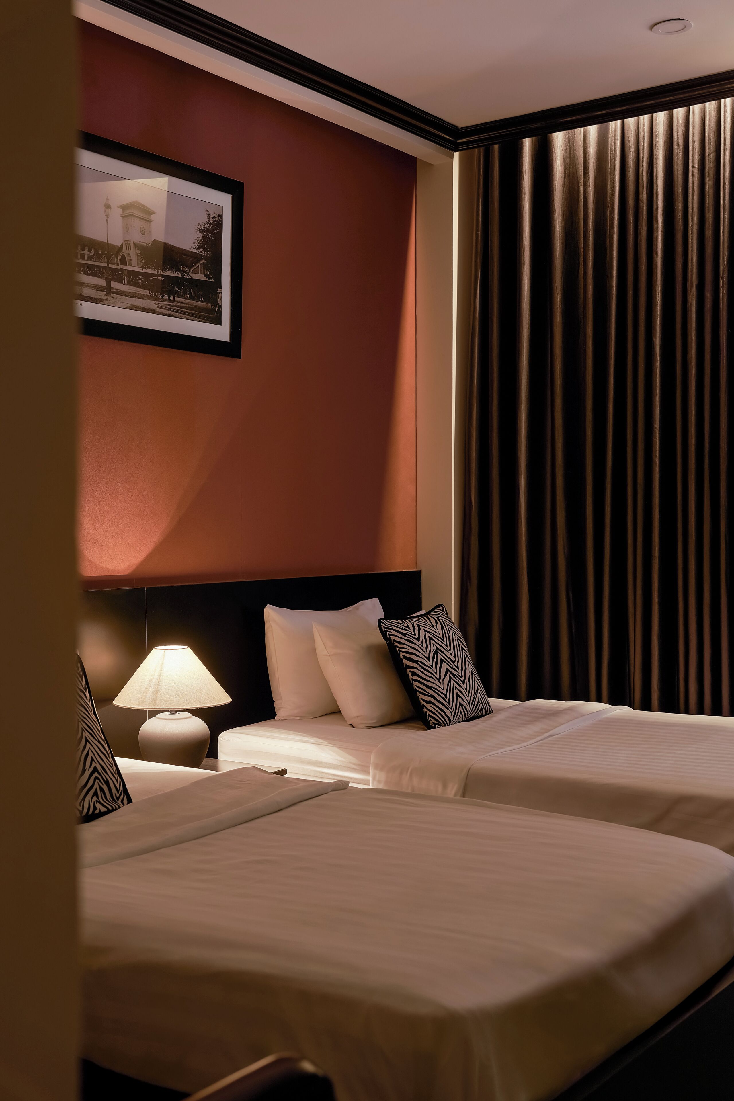 Habitación de hotel con dos camas, una mesita de noche con lámpara, un cuadro en la pared y un ventanal con cortinas.