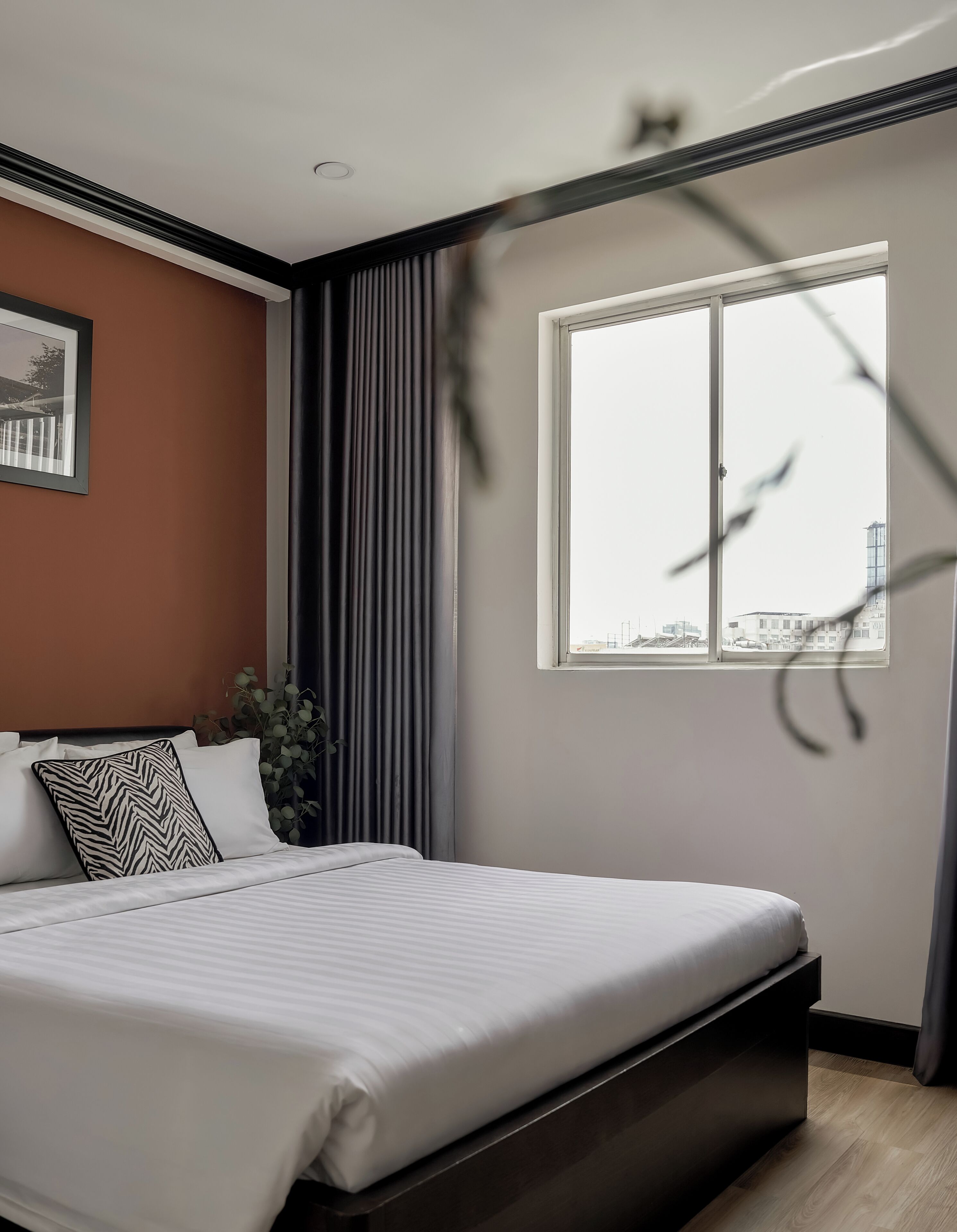 A modern bedroom with a large bed, a window with a city view, and a plant.