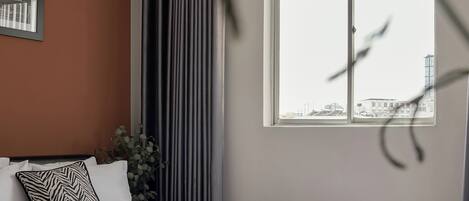 A modern bedroom with a large bed, a window with a city view, and a plant.