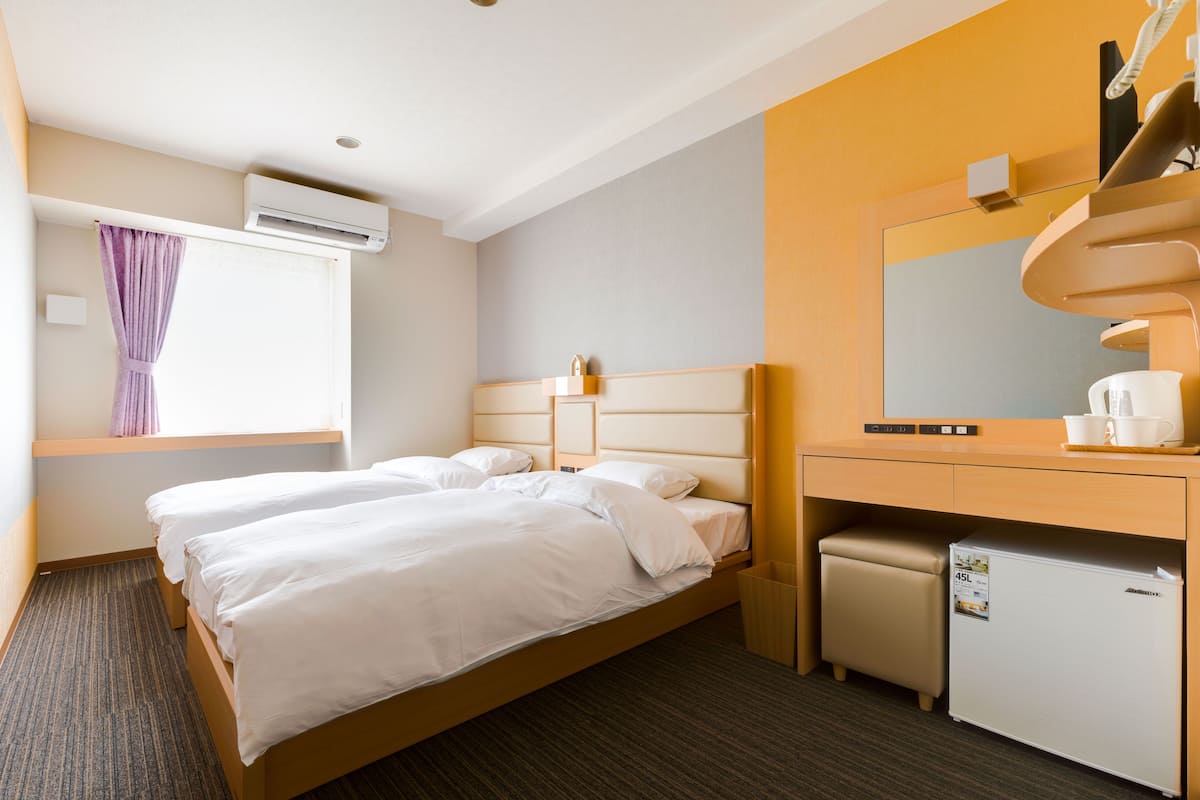 Quarto de hotel com duas camas, uma mesa de trabalho com espelho, um frigorífico pequeno e um aparelho de ar condicionado.