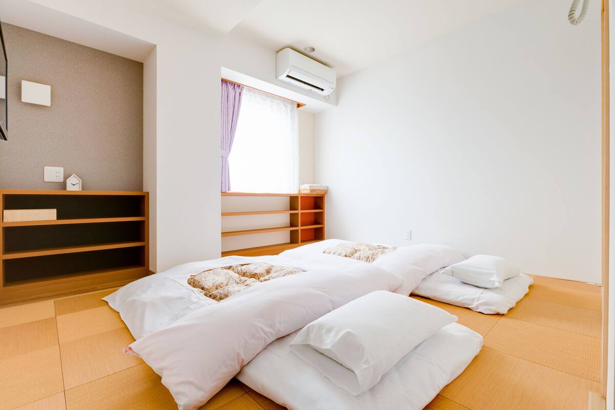 Quarto com duas camas, uma estante de madeira, janela com cortinas e aparelho de ar condicionado.
