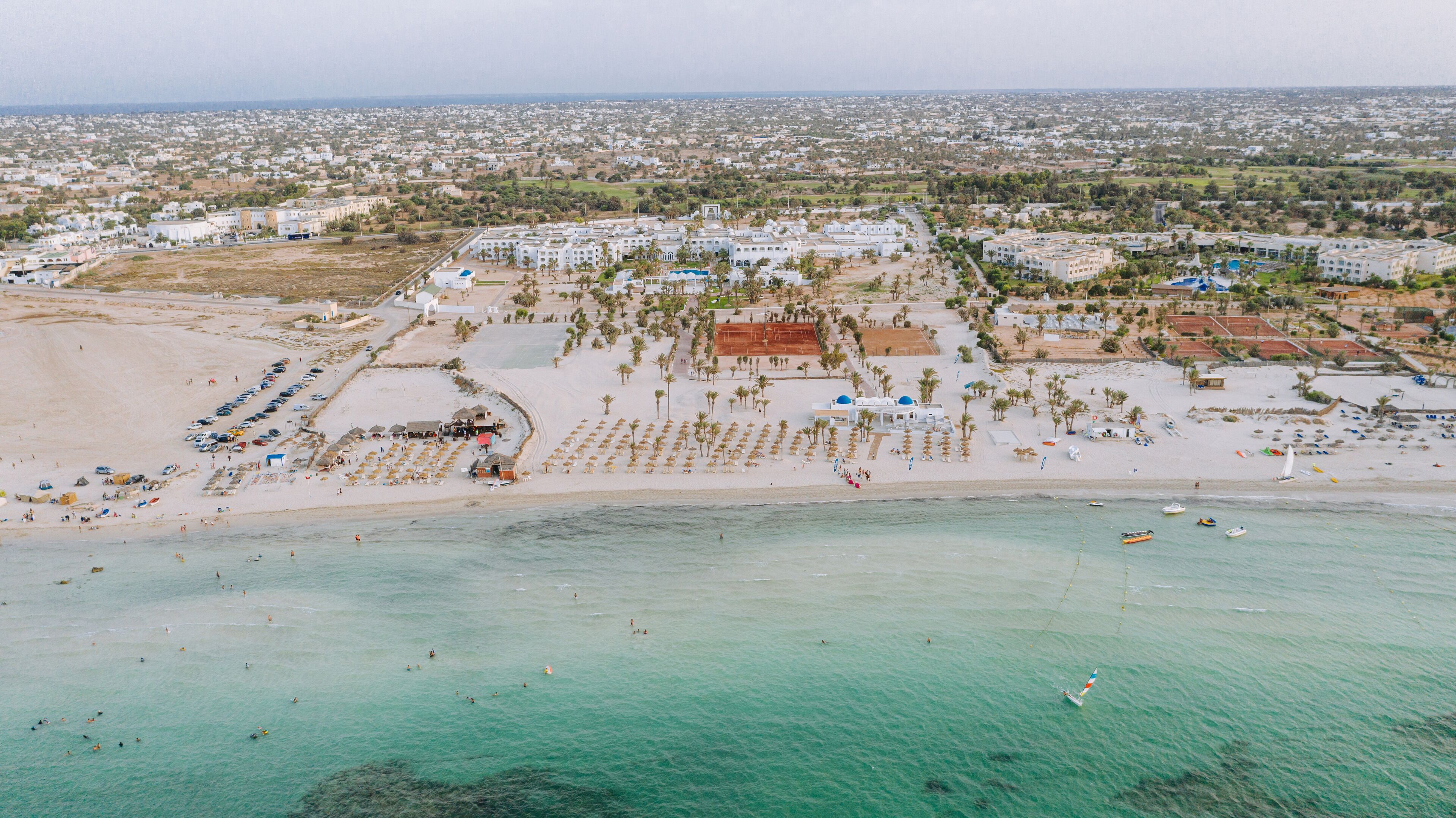 An aerial view of a coastal resort with multiple buildings, a sandy beach, and a clear turquoise sea.