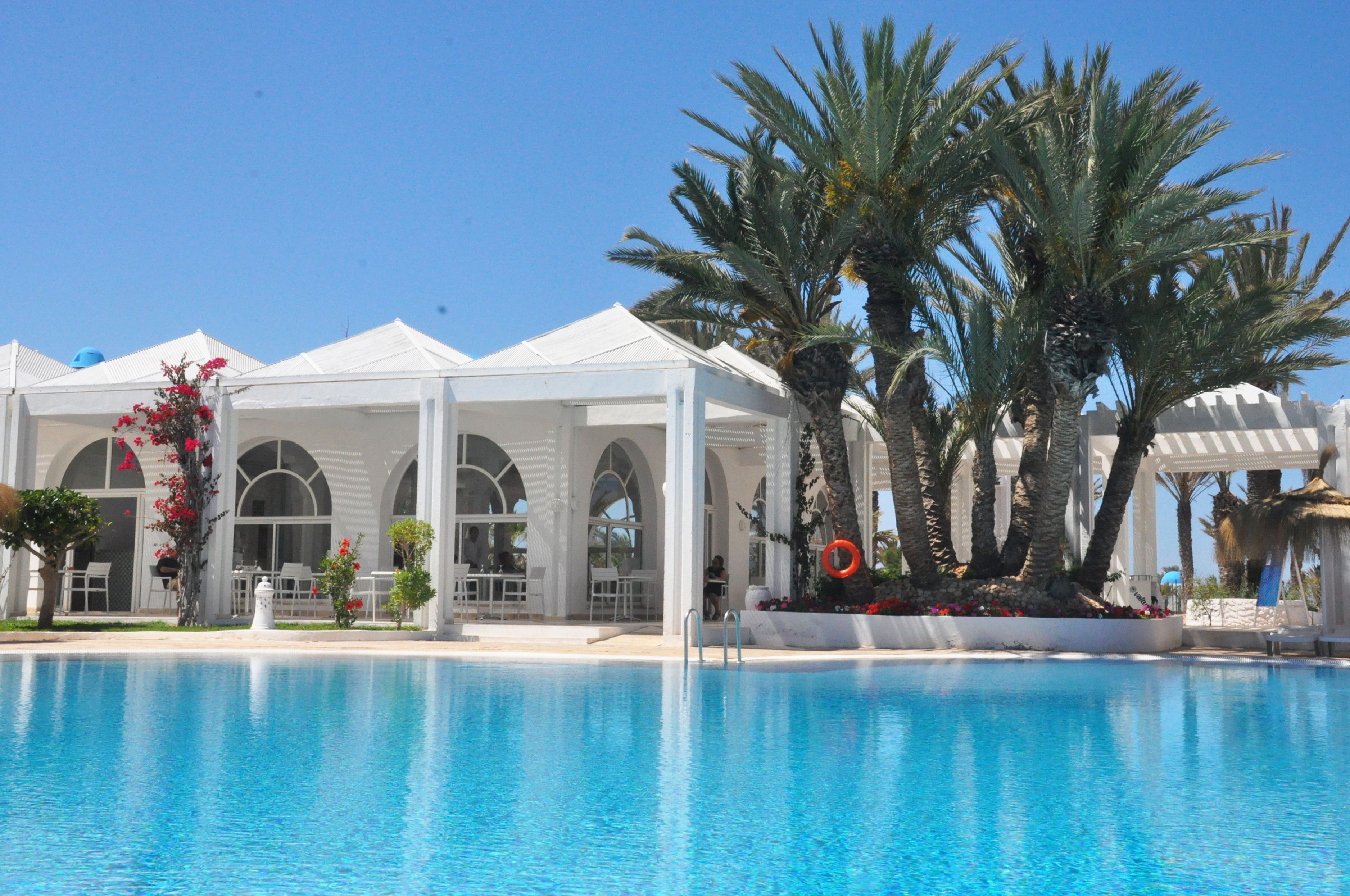 A poolside area with a clear blue pool, white canopied structures, palm trees, and a well-maintained garden.