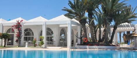 A poolside area with a clear blue pool, white canopied structures, palm trees, and a well-maintained garden.