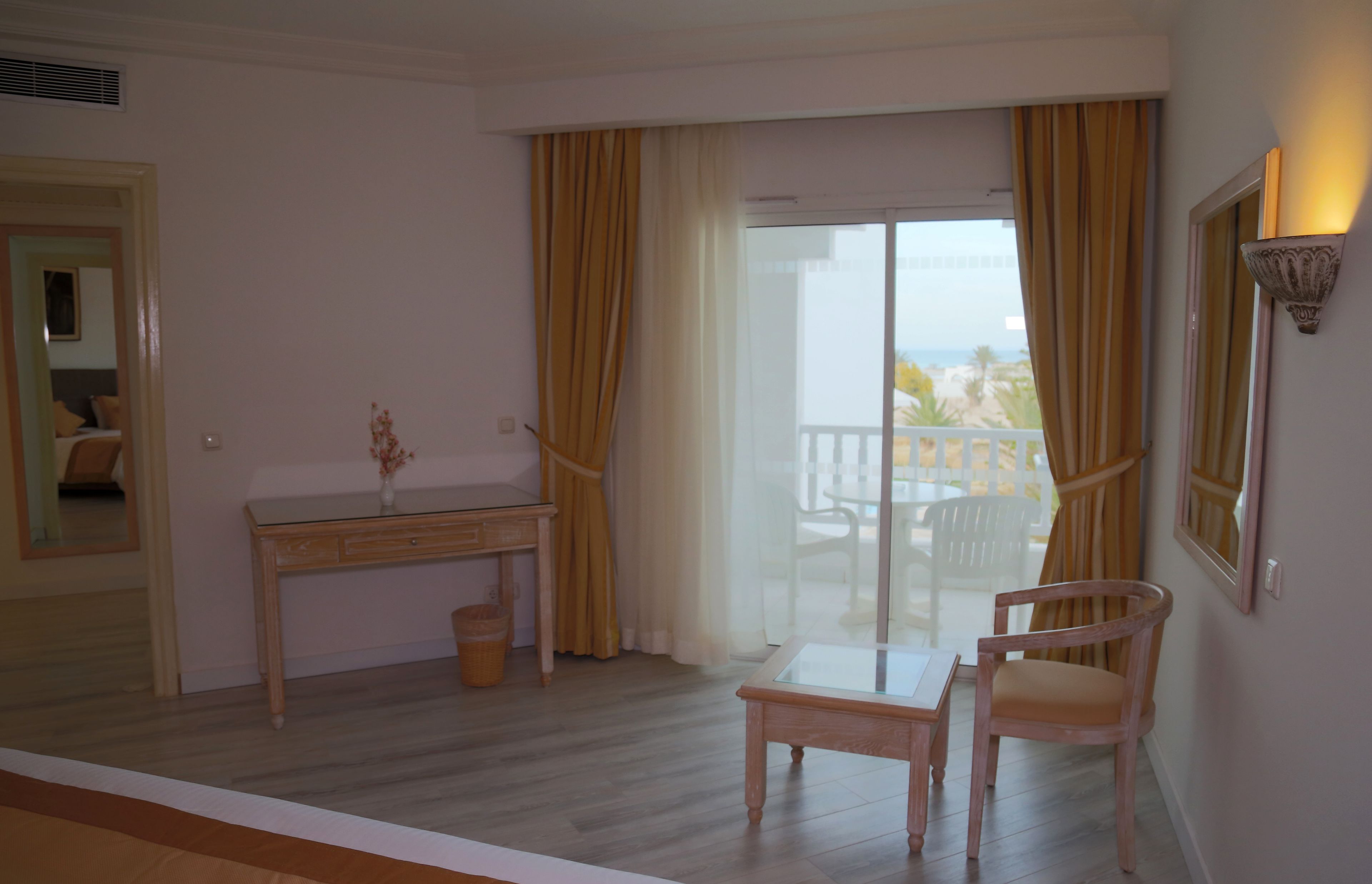 A hotel room with a wooden desk, a chair, a small table, a mirror, and a balcony with yellow curtains.