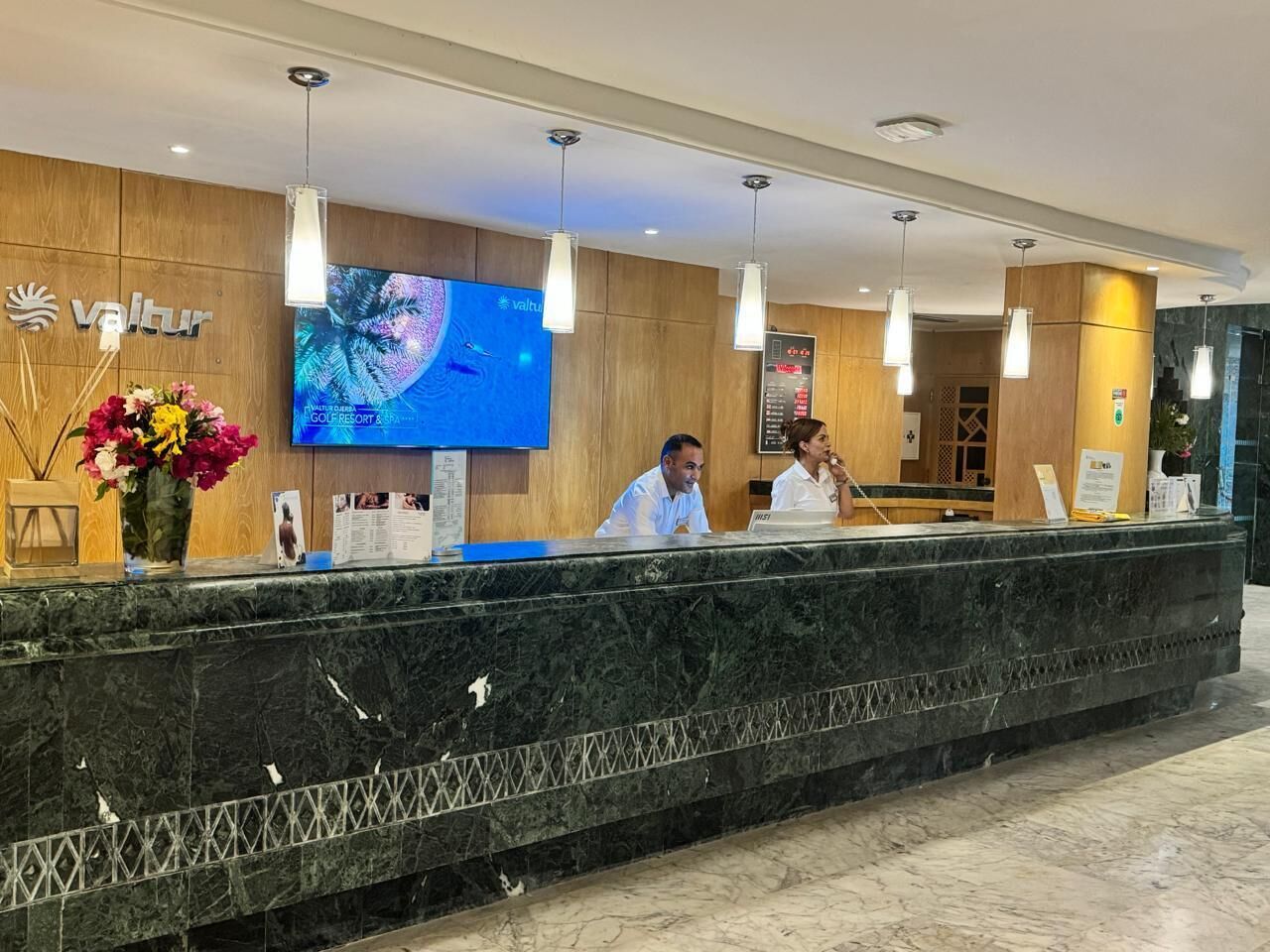 A hotel reception desk with a large screen displaying a tropical design, wooden paneling, and a marble counter.