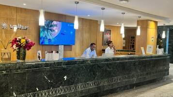 A hotel reception desk with a large screen displaying a tropical design, wooden paneling, and a marble counter.