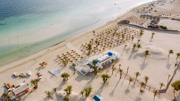 Vue aérienne d’un complexe hôtelier en bord de mer, avec une grande piscine, de nombreux parasols et des palmiers.