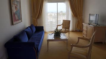 Una sala de estar con un sofá azul, una silla de madera, una mesa de centro de vidrio con un florero, un televisor sobre un mueble de madera, y un balcón con cortinas amarillas.