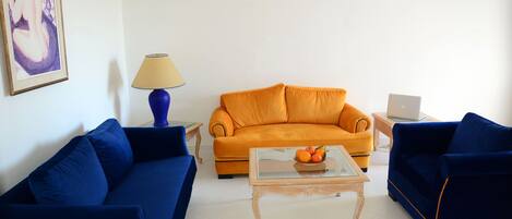 A living room with two sofas, one blue and one orange, a glass coffee table with fruit, a lamp, and a laptop on a side table.