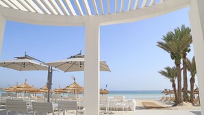 Une terrasse au bord de la plage, avec des chaises et des parasols blancs, des palmiers et une vue sur l’océan.