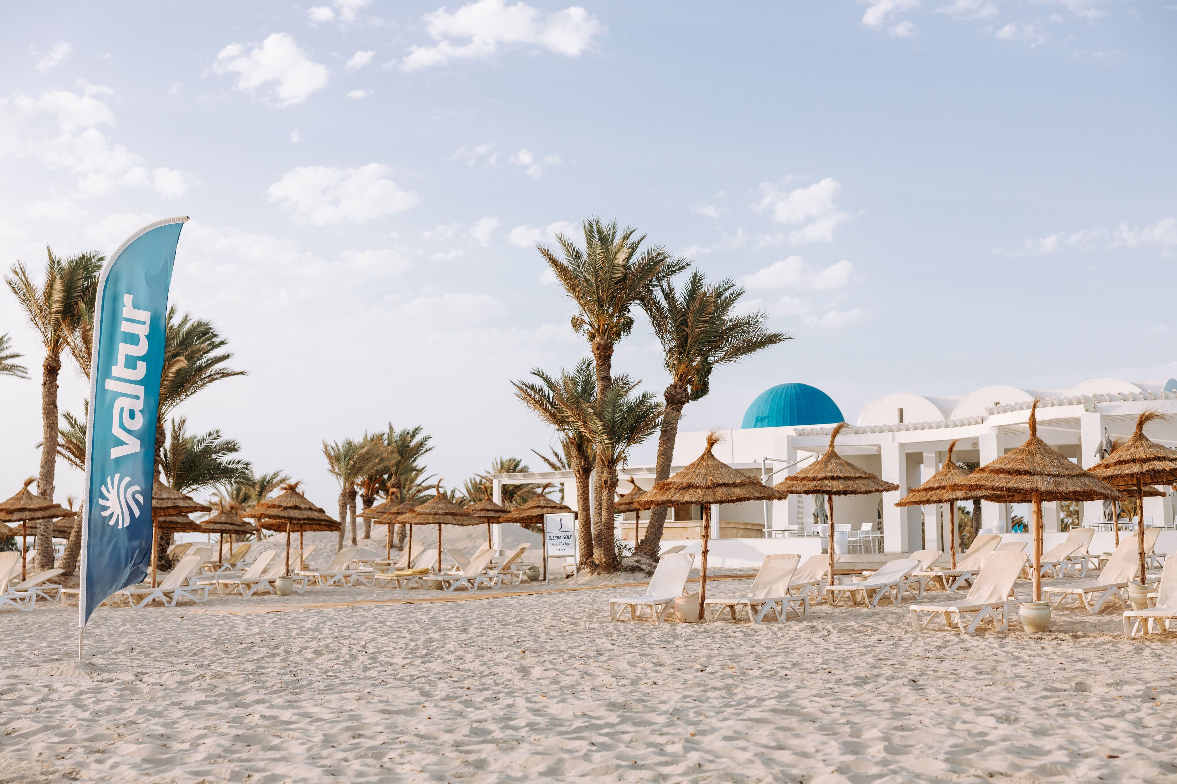 A beach with white lounge chairs, palm trees, and a blue flag with 'Valtur' written on it.