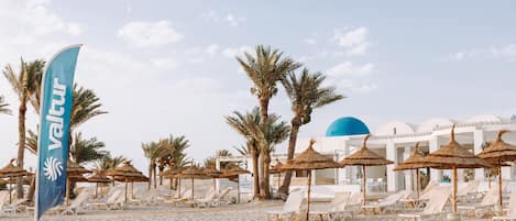 A beach with white lounge chairs, palm trees, and a blue flag with 'Valtur' written on it.