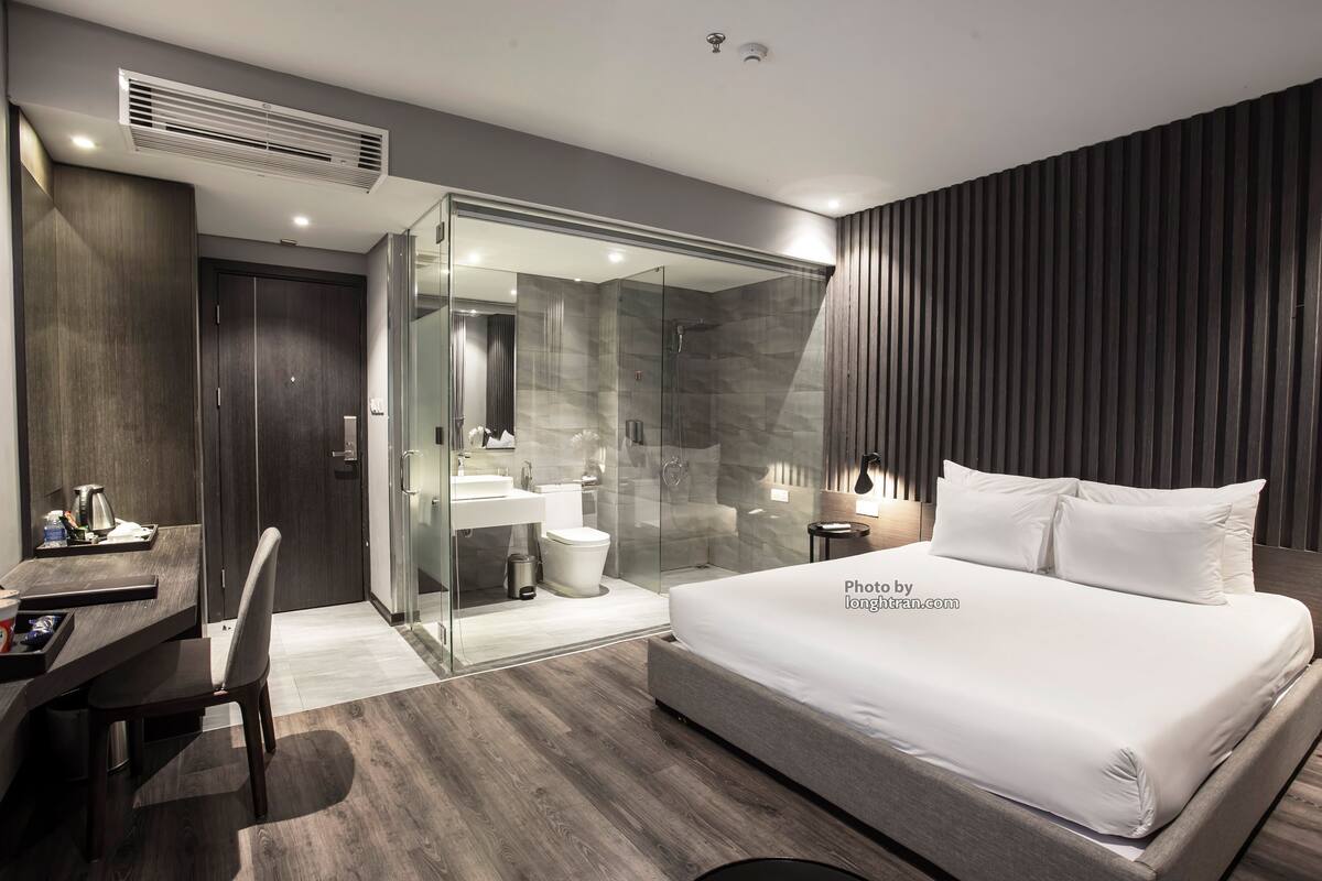 Habitación de hotel moderna con una cama grande, un escritorio, una silla, cabina de ducha con paredes de vidrio y un panel de pared de madera oscura.