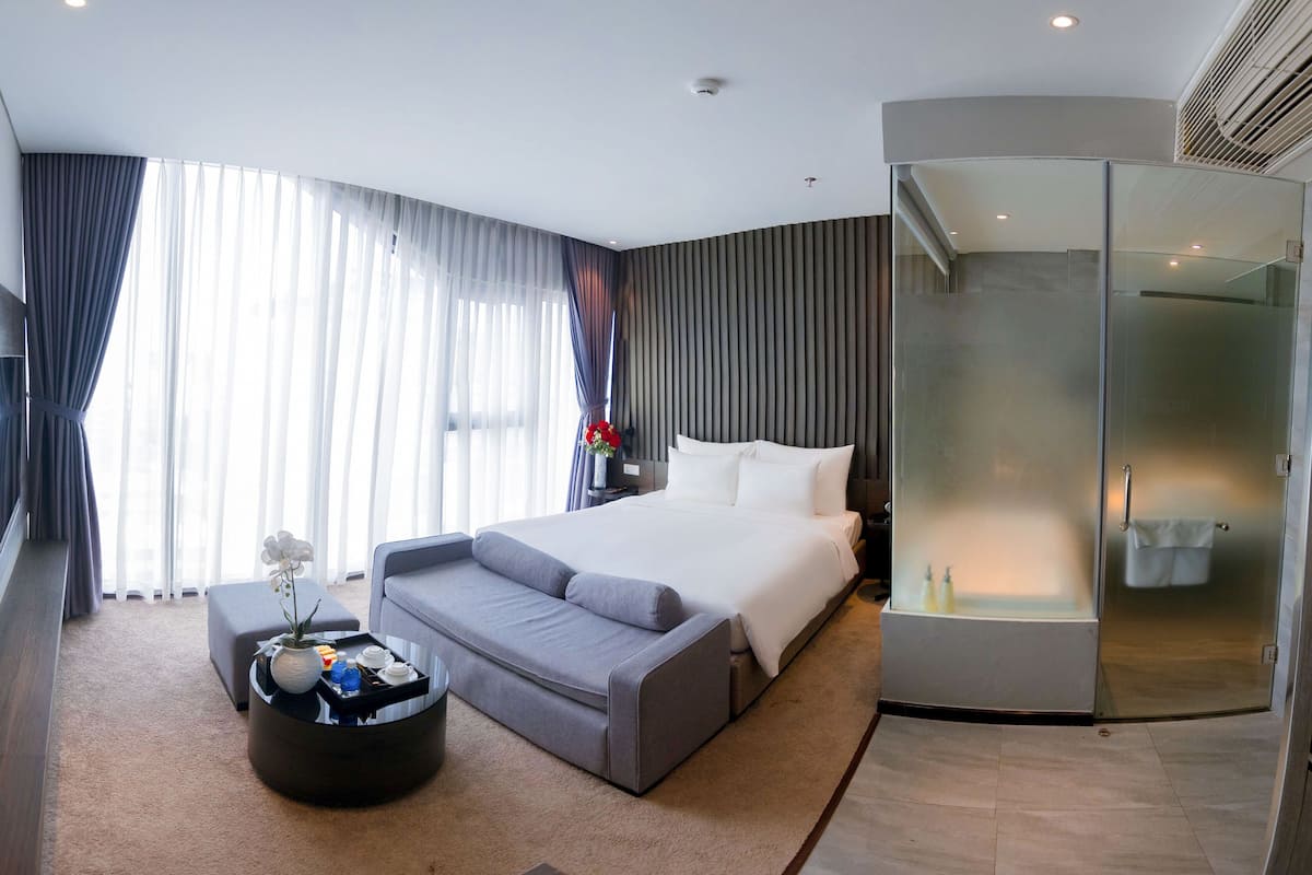 Una habitación de hotel moderna con una cama grande, una zona de estar con una mesa de centro, una ducha con paredes de vidrio y un ventanal con cortinas.