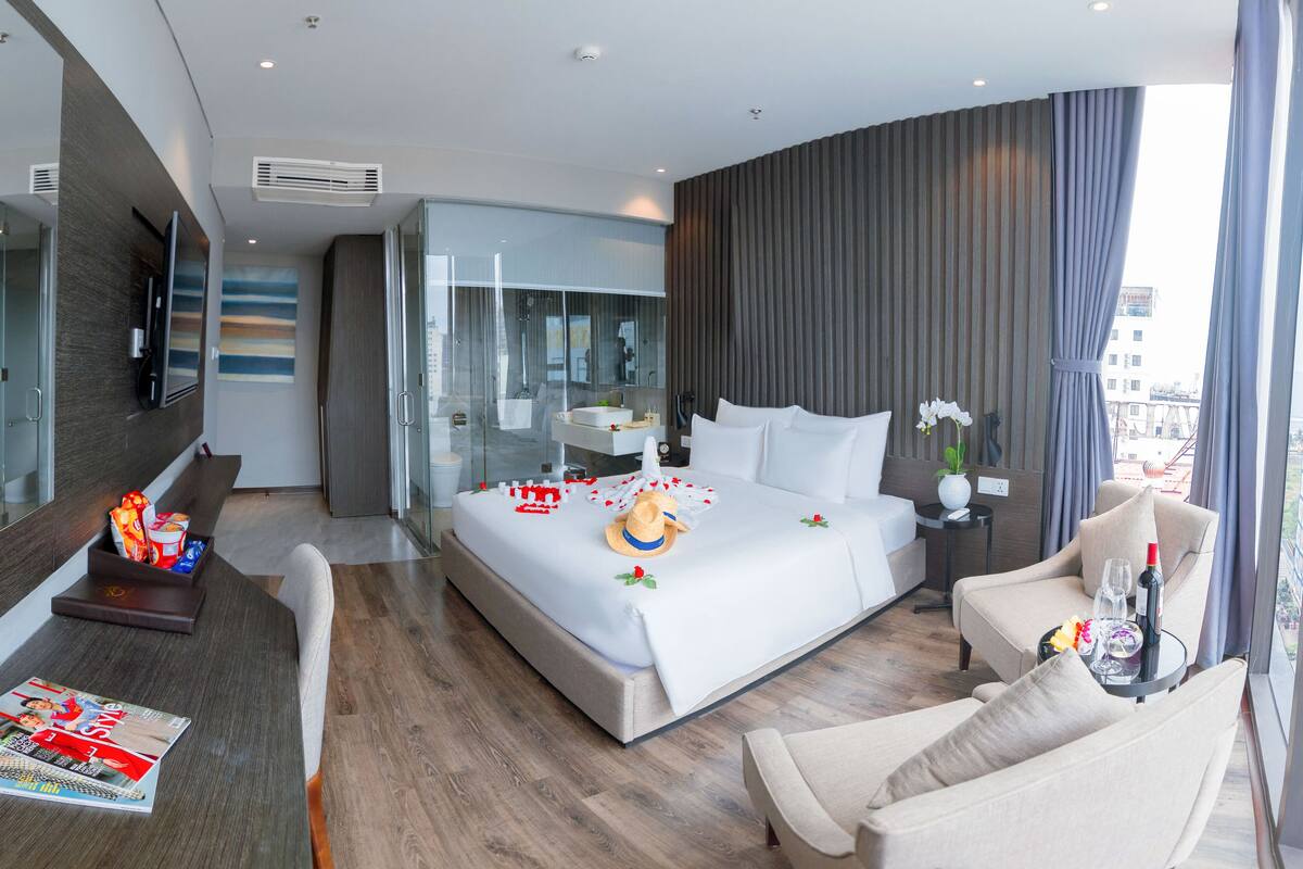 Una habitación de hotel moderna con una cama grande, una zona de estar con sofá y sillón, un televisor y un panel de vidrio que separa el dormitorio del baño.