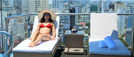 A balcony with two lounge chairs, one occupied by a person in a red bikini, and a cityscape view in the background.