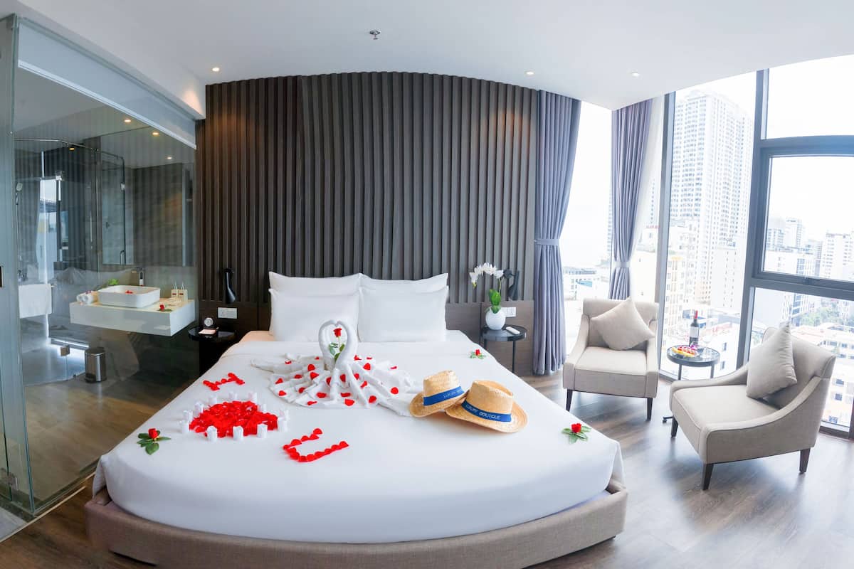 A modern hotel room with a large bed, a glass shower enclosure, a sitting area with a view, and a cityscape visible through the windows.