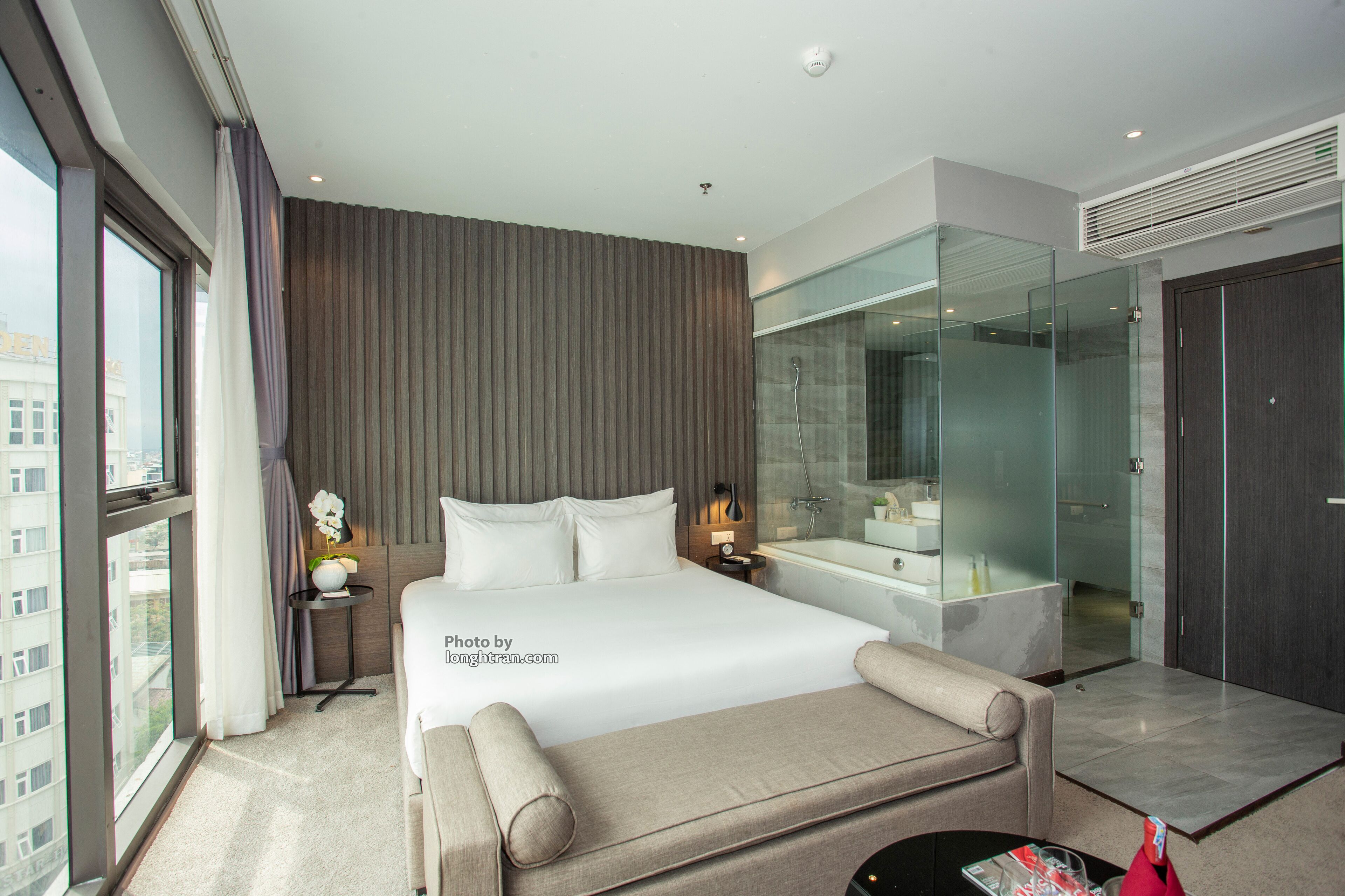 A modern hotel room with a large bed, a glass-enclosed shower, a sitting area, and a view of the city.