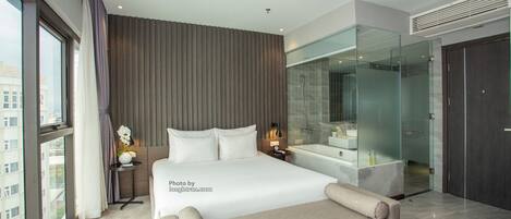 A modern hotel room with a large bed, a glass-enclosed shower, a sitting area, and a view of the city.